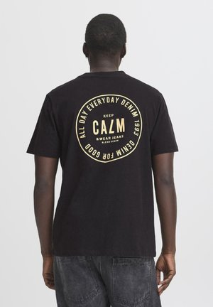 Maglietta nera in cotone con una grafica rotonda in testo dorato sul retro, che recita "KEEP CALM & WEAR JEANS," con una vestibilità casual e testurizzata.