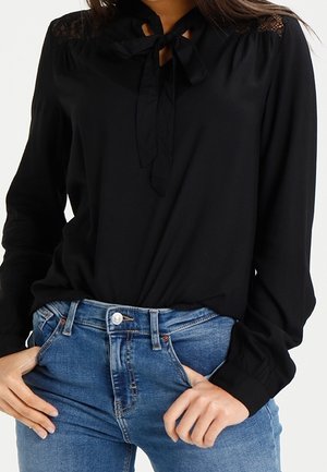 Femme portant un chemisier noir à manches longues avec un noeud sur le devant et des détails en dentelle, associé à un jean bleu taille haute.