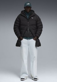 Sort pufferjakke med hætte, frontlynlås og elastiske manchetter, lagd over en grøn top. Parret med lysblå vide jeans og sorte sneakers.