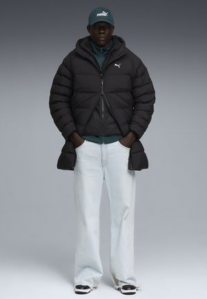 Giacca a vento nera con cappuccio, zip frontale e polsini elasticizzati, indossata sopra una maglietta verde. Abbinata a jeans a gamba larga blu chiaro e sneaker nere.