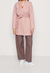 Cappotto rosa chiaro con cintura, colletto a scialle, maniche lunghe e tessuto texture, abbinato a pantaloni marroni larghi e sneakers bianche.