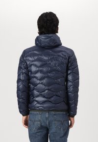 Giacca a vento blu navy con motivo trapuntato, finitura semilucida e colletto alto. Indossata sopra jeans blu, che mette in mostra una silhouette aderente.