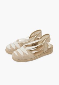 Béžové espadrilky s křížovými bílými plátěnými popruhy, otevřenou zadní částí a klínovým podpatek omotané jutou, s texturovaným tkaninovým materiálem.