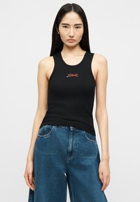 Νεαρή γυναίκα με μακριά μαύρα μαλλιά φοράει μαύρο αμάνικο τοπ με μικρό γραφικό τίγρης και άνετο μπλε τζιν, στέκεται μπροστά σε λευκό φόντο.