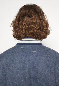 Blå ulljacka med en ribbad marinblå och vit krage, med vita sömnadsdetaljer och en avslappnad passform bak.