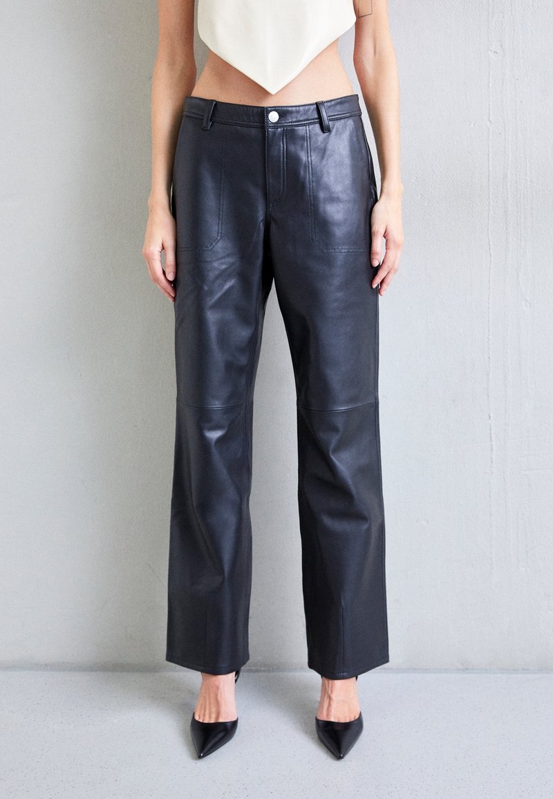 Femme portant un pantalon en cuir noir taille haute, un top court asymétrique blanc, et des talons noirs à bouts pointus, debout contre un mur gris.