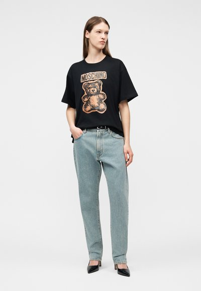 Γυναίκα στέκεται φορώντας φαρδύ μαύρο T-shirt με καφέ αρκουδάκι και την επιγραφή "MOSCHINO", ανοιχτόχρωμα μπλε τζιν, μαύρες ψηλοτάκουνες, με τα χέρια στις τσέπες.