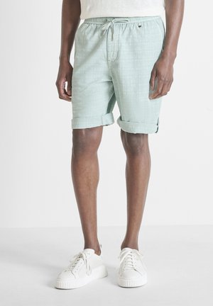 Uomo con pantaloncini verde chiaro risvoltati con coulisse e sneaker bianche, in piedi su uno sfondo chiaro e uniforme.