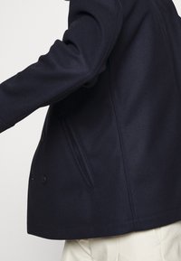 Chaqueta marinera de corte cruzado, hecha de una tela texturizada, con un bolsillo lateral y una silueta estructurada, con detalles de costura visibles.