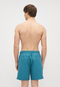 Tyrkysové plavky vyrobené z ľahkého materiálu, s elastickým pásom, zadnou vreckom a hladkým povrchom. Zadný pohľad na model.