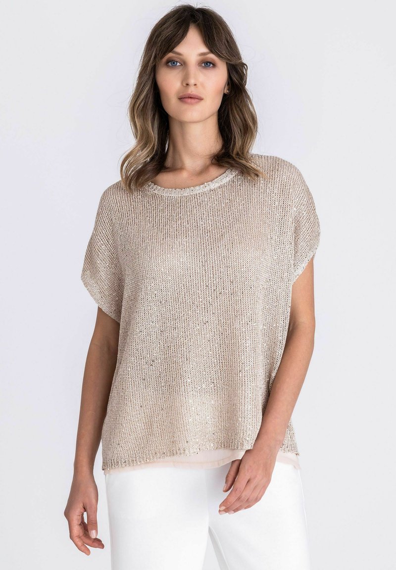 Vrouw met schouderlang haar, gekleed in een losse beige gebreide top over een lichte blouse en witte broek tegen een effen achtergrond.