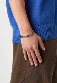 Braccialetto in argento con forme geometriche intrecciate, indossato su un polso, accanto a un maglione blu tessurizzato e pantaloni marroni.
