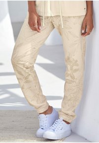 Beige Sweatpants mit floralem Muster, elastischen Bündchen und einem Kordelzug in der Taille, kombiniert mit weißen Sneakers. Glatte Stofftextur.