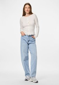 Mujer joven con cabello largo que lleva un cárdigan blanco abotonado, unos vaqueros de pierna ancha en azul claro y zapatillas plateadas, de pie con las manos en los bolsillos.