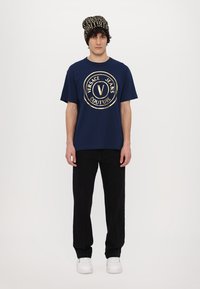Tumši zils t-krekls ar zelta apļu logo, uz kura rakstīts "Versace Jeans Couture", apvienots ar melniem biksēm un rakstainu adītu cepuri.