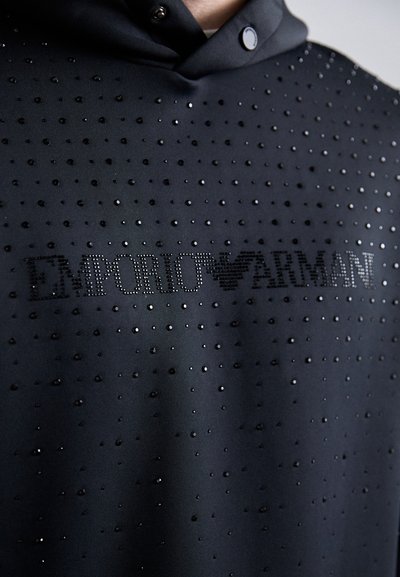 Hoodie preto com uma textura suave, apresentando um padrão de pequenos studs brilhantes e o logótipo "EMPORIO ARMANI" em embelezamentos semelhantes a cristais.