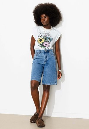 Femme avec une coiffure afro portant un t-shirt blanc à motif floral, un short en jean bleu au genou et des chaussures marron à enfiler sur fond blanc.