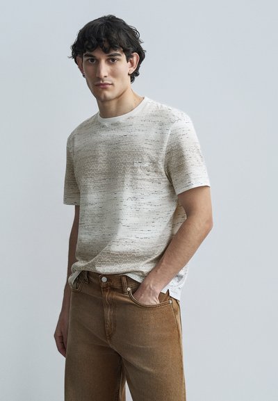 Short-sleeved beige T-shirt with a textured zigzag pattern, featuring a small logo on the chest, paired with brown denim jeans.
