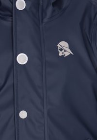 Navy zila ūdensizturīga jaka ar gludu tekstūru, kurai ir lieli balti pogas ar snapu un sudraba logotips ar sejas profilu.