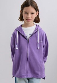 Fioletowa bluza na suwak z kapturem ze sznurkiem, przednimi kieszeniami i haftowanym wzorem. Gładki materiał o luźnym kroju, noszona na białym t-shircie.