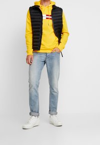 Hombre con sudadera amarilla de Tommy Hilfiger, chaleco acolchado negro, jeans ligeros arremangados y zapatillas blancas frente a un fondo blanco liso.