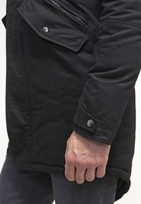 Svart, vadderad jacka med en zippered ficka, knäppt ärmslut och en slät textur, med en något förlängd nederkant.