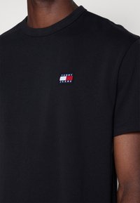 Czarny bawełniany t-shirt z okrągłym dekoltem, z małym wielokolorowym haftowanym logo na lewej stronie klatki piersiowej. Krótkie rękawy i gładka faktura.