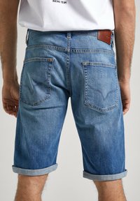 Shorts de mezclilla en azul medio, con dobladillos enrollados, dos bolsillos traseros con detalles de costura y un parche de cuero en la cintura.