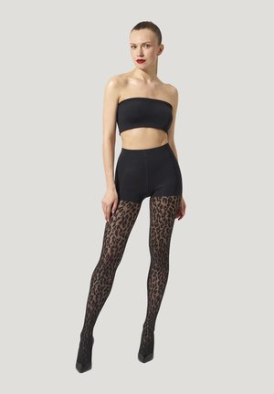 Donna che indossa un crop top nero senza spalline, pantaloni corti neri, collant trasparenti con stampa leopardata e tacchi a punta neri, in piedi davanti a uno sfondo semplice.
