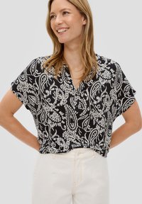 Zwarte kortemouwtop met een wit paisleypatroon, kraag, en lichtgewicht stof. Gecombineerd met lichtgekleurde jeans.