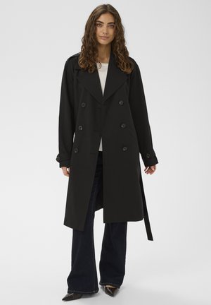 Fekete, dupla soros esőkabát nagy gombokkal, széles hajtókával és állítható mandzsettákkal; fehér felső fölött viselve, sötét farmerral párosítva.