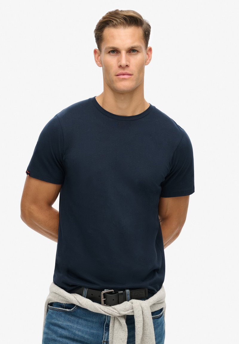 Dunkelblaue Baumwoll-T-Shirt mit kurzen Ärmeln und Rundhalsausschnitt; verfügt über ein kleines Logodetail am Ärmel und wird mit hellblauen Jeans kombiniert.