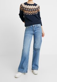 Pull marin avec motif fair isle en blanc et marron, associé à un jean large bleu clair et des baskets blanches. Tenue décontractée.