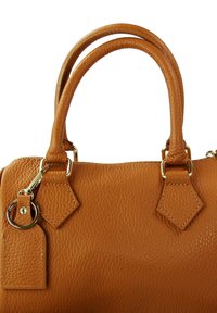 Tan leren handtas met textuur aan de buitenkant, twee ronde handvaten, diamanten accenten en een sleutelhanger van goudkleurig metaal.