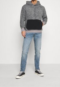 Felpa grigia in pile con tasca frontale nera, dettagli in grigio chiaro ai polsini, abbinata a jeans di denim azzurri e sneakers nere.