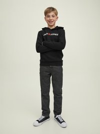 Felpa con cappuccio nera e logo bianco, abbinata a jeans grigi scuri e sneakers nere con dettagli bianchi. Il modello è in piedi con le braccia incrociate.