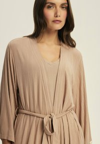 Beżowy szlafrok z prążkowanym wzorem, posiadający dekolt w kształcie V, szerokie rękawy i przewiązywany pasek. Teksturowany materiał tworzy subtelne linie i luźny krój.