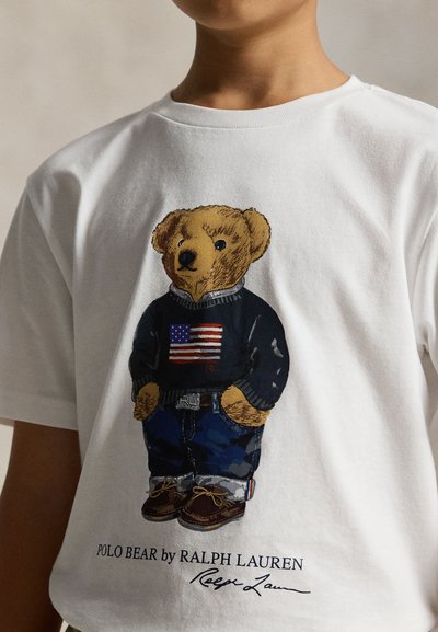 Child wearing white shirt with graphic of teddy bear dressed in jeans, navy sweater with American flag, and brown shoes.