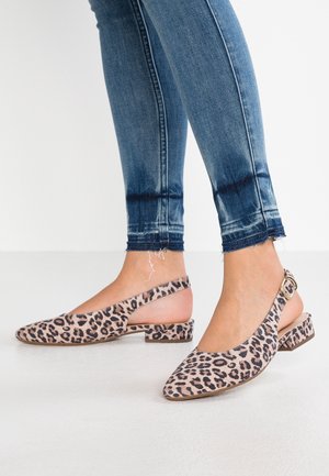 Pieds portant un jean skinny bleu et des ballerines à bride arrière à imprimé léopard, à talon bas et boucle, sur un sol blanc.
