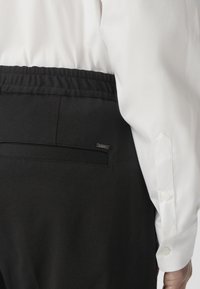 Zwarte broek met een elastische tailleband, voorzien van een achterzak en een klein metalen logo-label. Witte shirt met knoopmanchetten zichtbaar.