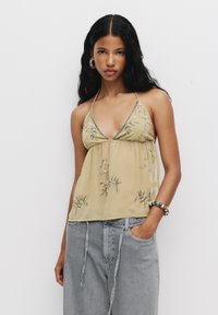 Top halter cu dantelă în galben deschis, cu broderie argintie, material transparent și croială lejeră, purtat cu pantaloni gri și o brățară mare.