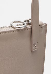 Bolso de cuero color topo claro con cremallera, herrajes plateados y un detalle de costura en el asa. Textura lisa, diseño minimalista.