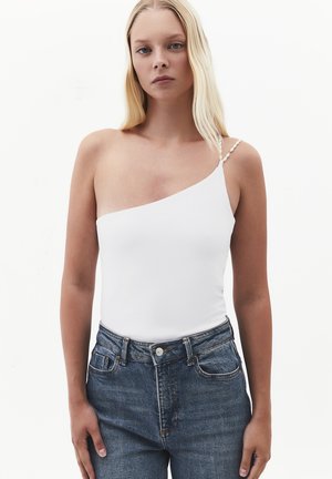 Joven mujer con cabello rubio largo que lleva una camiseta blanca de un solo hombro y vaqueros azules de cintura alta sobre un fondo blanco liso.
