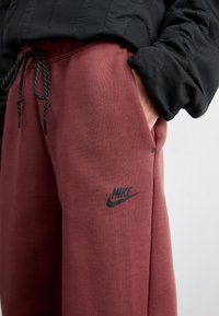 Calças de treino Nike em tecido bordô, com bolsos laterais, um cordão na cintura e um logótipo preto na coxa esquerda. Textura suave.