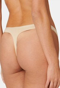 Beige Tanga-Unterwäsche mit glatter Textur, minimaler Bedeckung und niedrigem Bund, ausgestattet mit einem einfachen elastischen Bund.