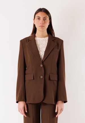 Donna con capelli lunghi e scuri che indossa una giacca marrone sopra una maglia bianca di pizzo, in piedi davanti a uno sfondo chiaro uniforme, rivolta verso l'obiettivo.