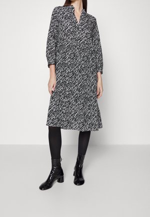 Vrouw die een zwart-witte midi-jurk met lange mouwen en abstract patroon draagt, zwarte panty's en glanzende zwarte enkellaarsjes, staand tegen een effen achtergrond.