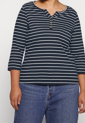 Personne portant un t-shirt à manches longues à rayures horizontales bleu marine et blanches avec quatre boutons au niveau de l'encolure et un jean en denim bleu.