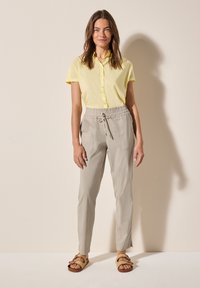 Camicia button-up gialla chiara abbinata a pantaloni grigi chiari con vita elastica e tasche laterali, e sandali marroni con cinturini. Texture del tessuto morbida.