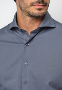 Camicia grigia con colletto allargato, chiusura a cinque bottoni e texture di tessuto liscia. I dettagli distintivi includono colori dei bottoni a contrasto.